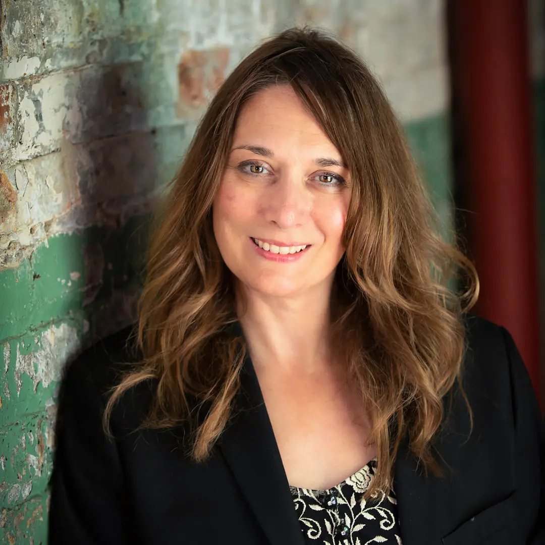 Head shot of Visionary Artist Lisa Nomikos leaning against a white and green brick wall at Starline Art Gallery in Harvard, Illinois.